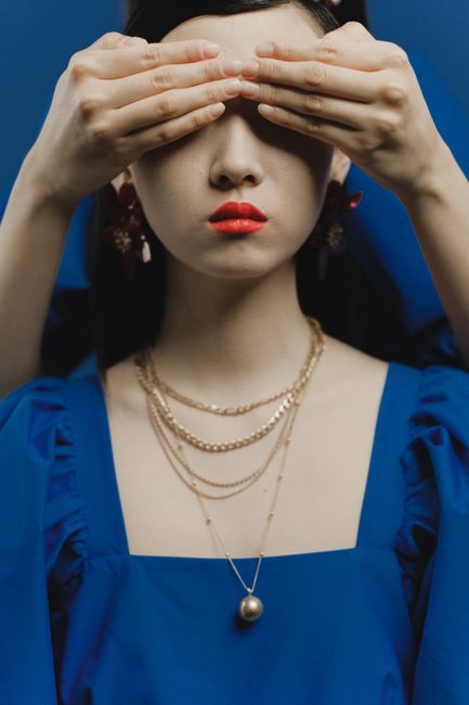 A woman wears layered gold necklaces and earrings with red lips, as hands cover her eyes.