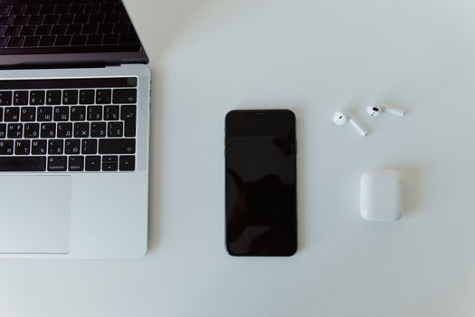 Top view of a laptop, smartphone, and wireless earphones on a white desk.