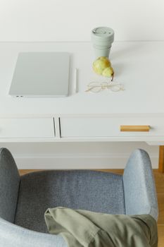 A clean and minimalist workspace with a laptop, pear, and reusable cup on a white desk.