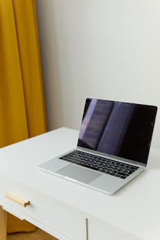 Elegant silver laptop on a simple white desk with mustard curtain backdrop.