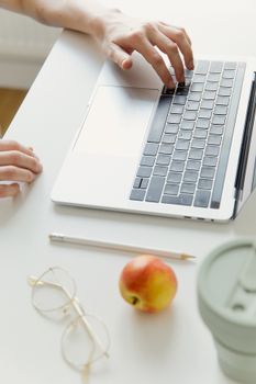 Close-up of hands typing on a laptop beside an apple and eyeglasses on a white desk.