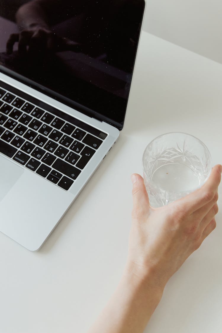 A Hand Holding A Glass Of Water Beside Laptop On A White Surface