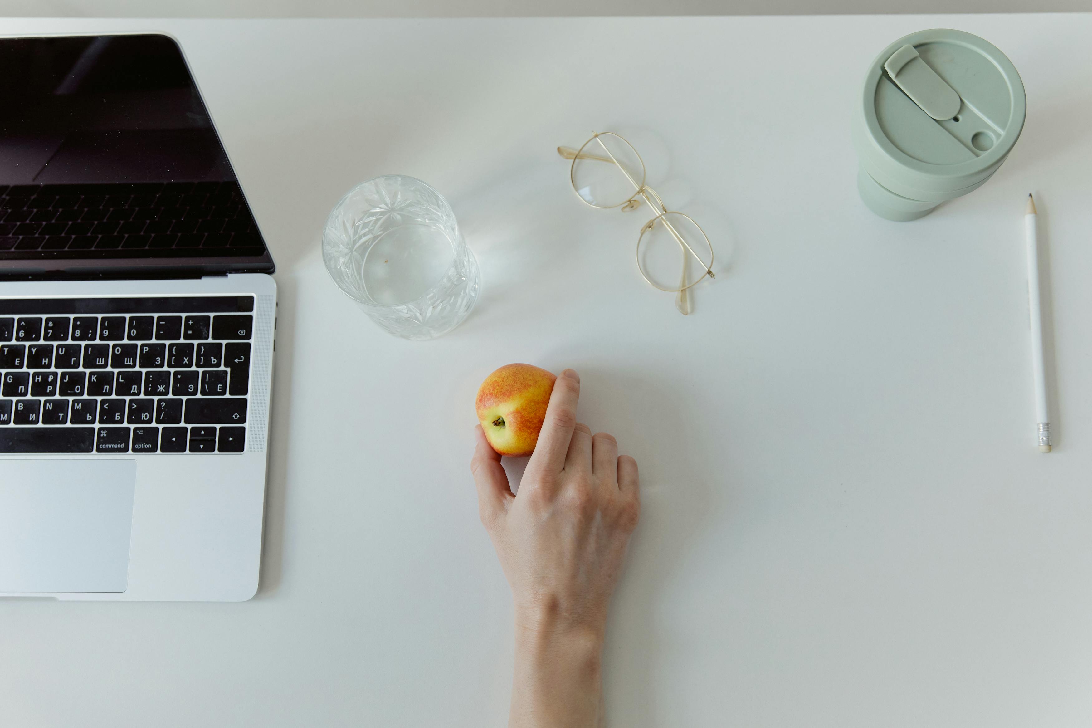 Overhead view of a minimalist workspace featuring a laptop, apple, glass, and eyeglasses on a white surface.