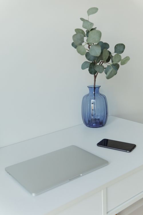Free Simple workspace setup with a laptop and smartphone beside a blue vase with greenery on a white table. Stock Photo