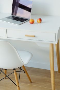 Modern minimalist workspace featuring a sleek laptop and two fresh apples on a white desk with a white chair.