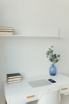 A minimalist white workspace featuring a laptop, phone, books, and a vase with eucalyptus on a white desk.