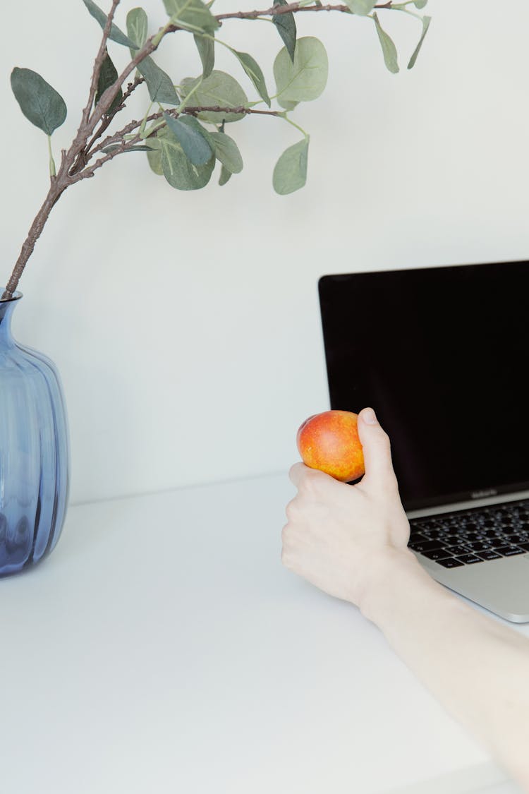 A Hand Holding An Apple Beside Laptop On A White Surface