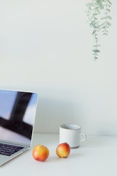 A bright and minimalist workspace featuring a laptop, mug, and fresh fruit on a white surface.