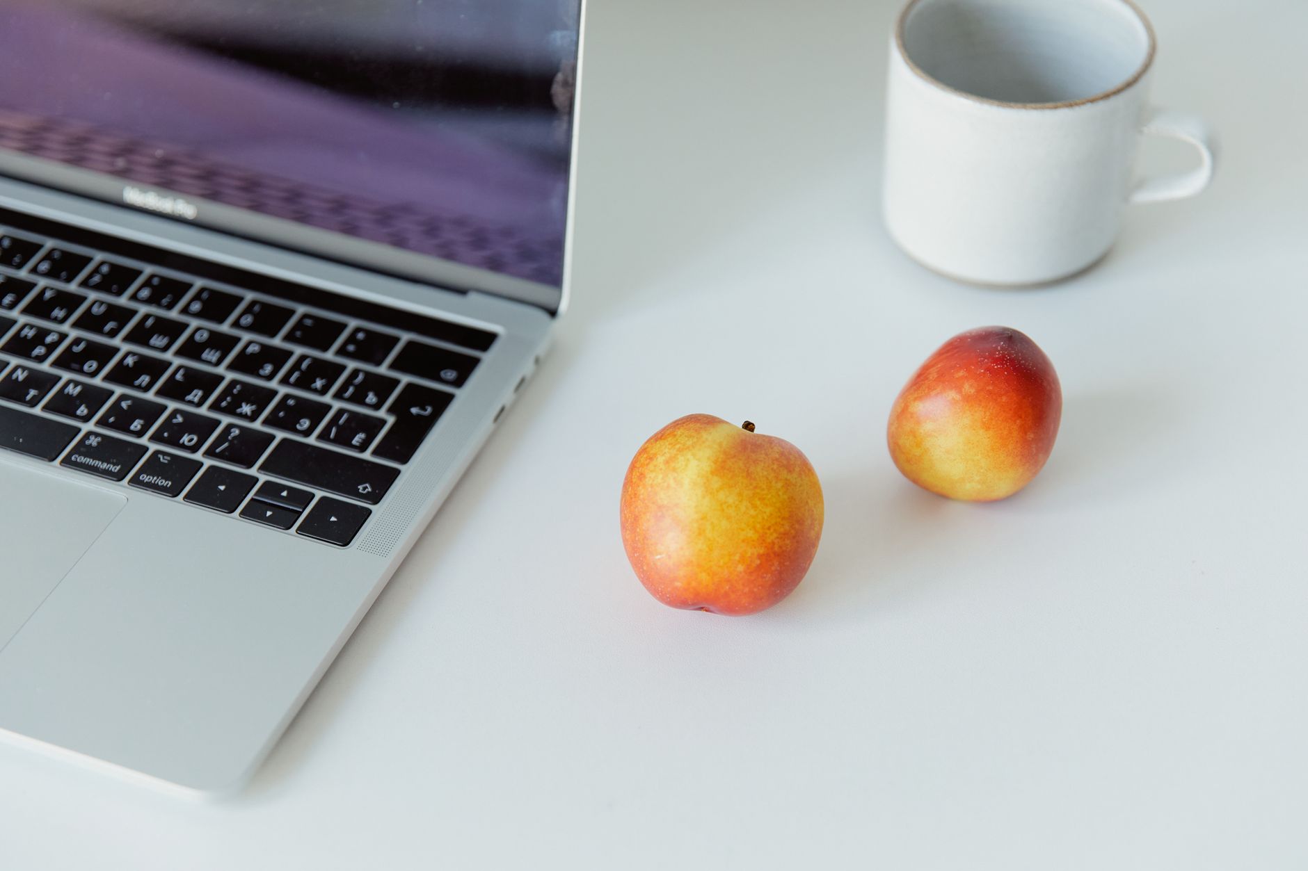 Clean desktop with laptop, apples, and mug on a white surface. Ideal for workspaces.