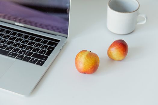 Clean desktop with laptop, apples, and mug on a white surface. Ideal for workspaces.