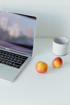 A minimalistic work desk featuring a laptop, fresh apples and a cup on a white surface.