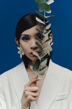 Portrait of a stylish woman with bold eyeshadow holding eucalyptus against a blue background.