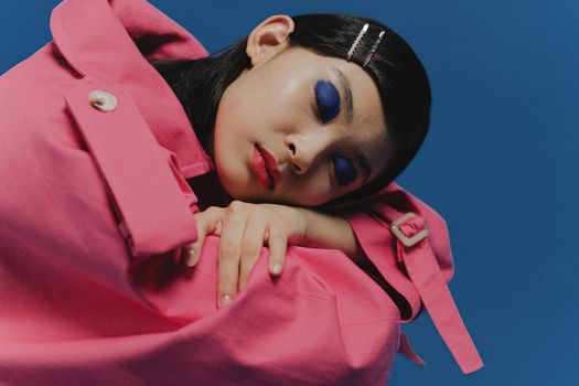 Portrait of an Asian woman in a pink coat with blue eyeshadow against a blue background.