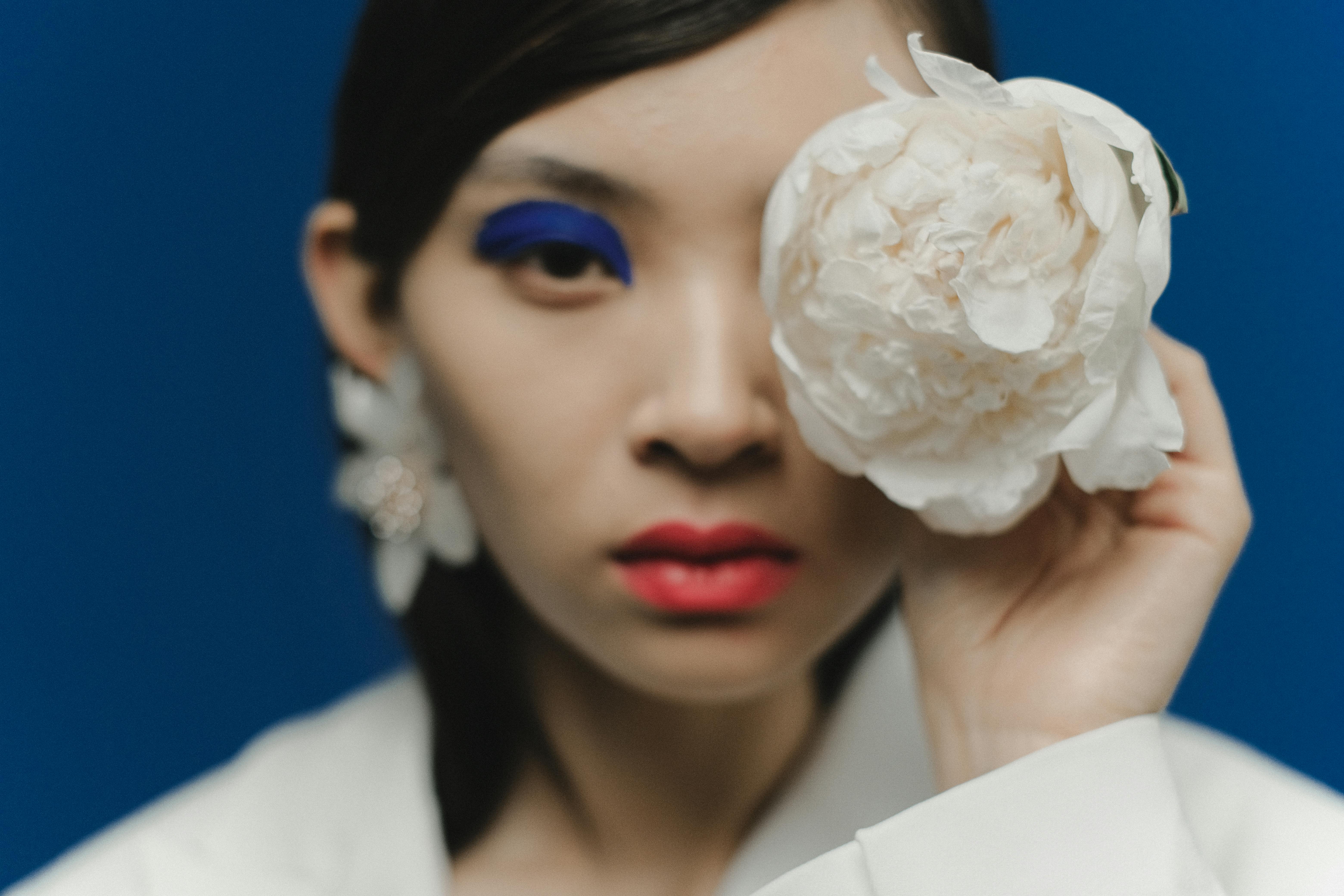 Stylish portrait of a woman with blue makeup holding a white flower against a blue background.