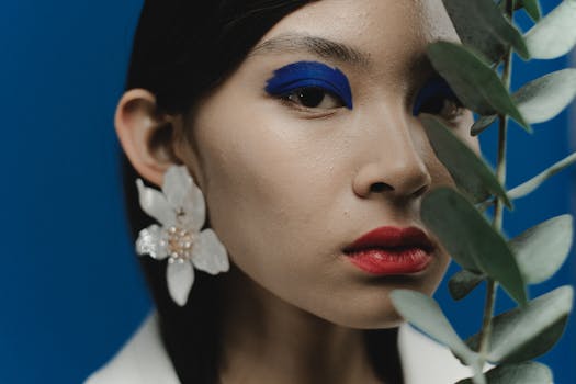 Close-up portrait of a woman with vibrant makeup and floral earrings, framed by greenery.