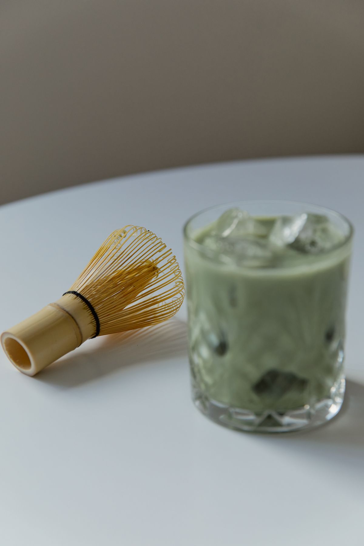Pale green iced matcha latte in a cut glass tumbler next to a bamboo whisk