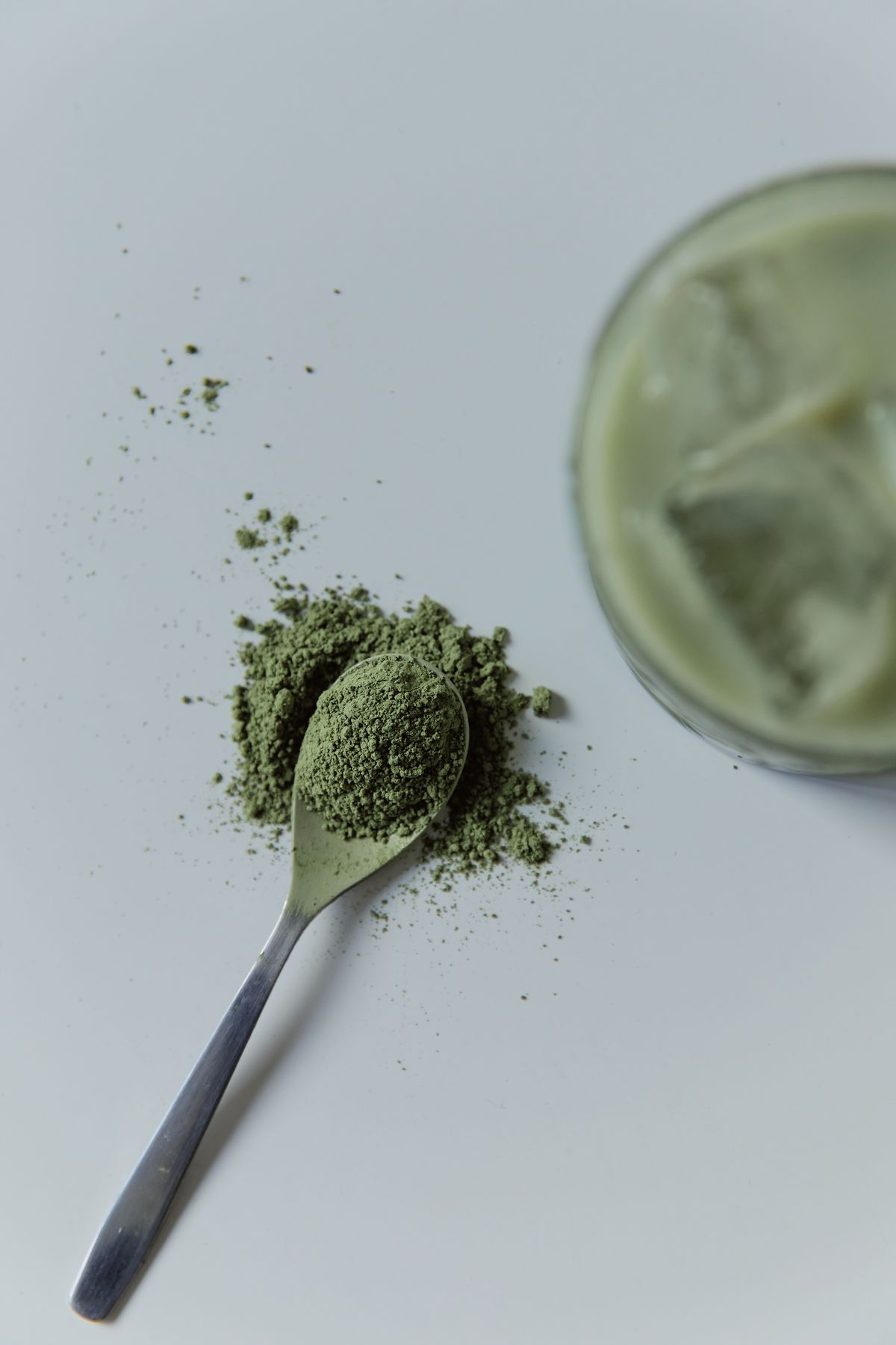 Green tea powder on a metal spoon beside a pale iced drink on a white surface