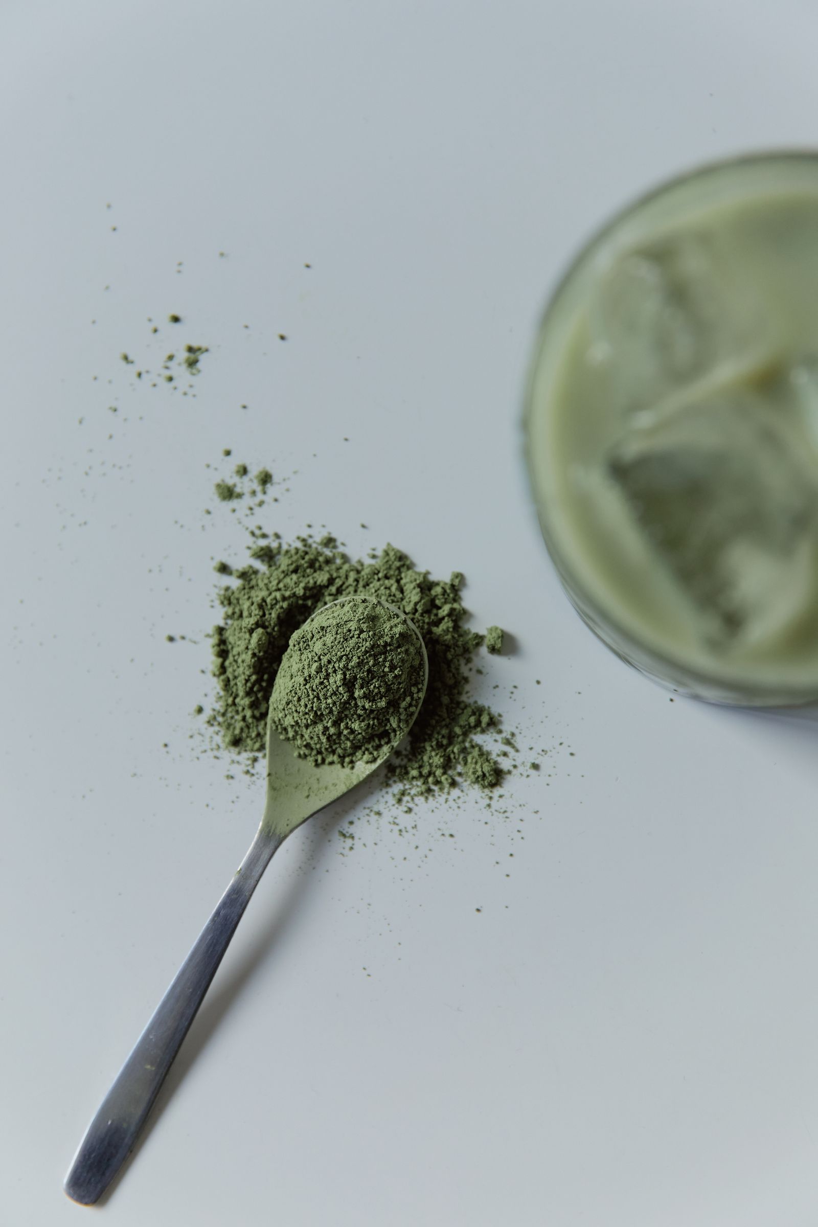 A spoon heaped with bright green matcha powder on a white surface next to a glass of iced matcha