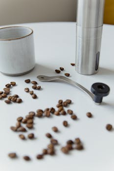 A minimalist coffee setup with beans, grinder, and cup on a white table.
