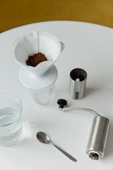 Overhead view of coffee brewing tools including a dripper, grinder, and spoon on a white table.