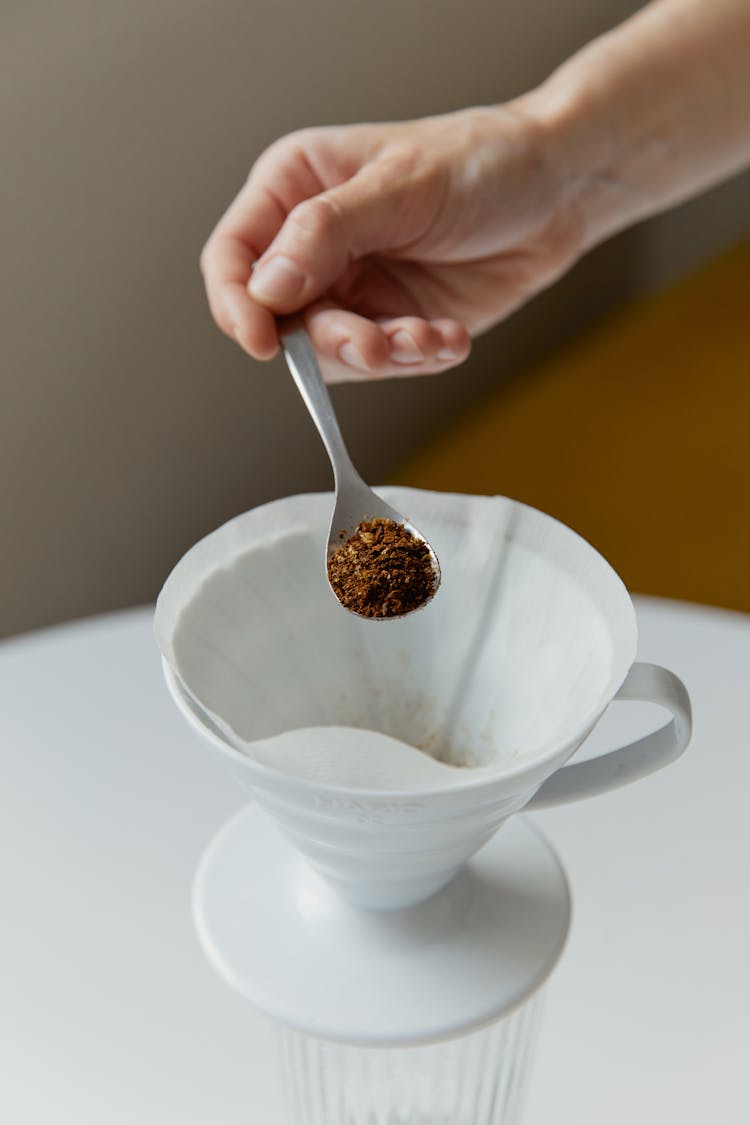 A Hand Holding A Teaspoon Of Coffee Powder Near White Cup