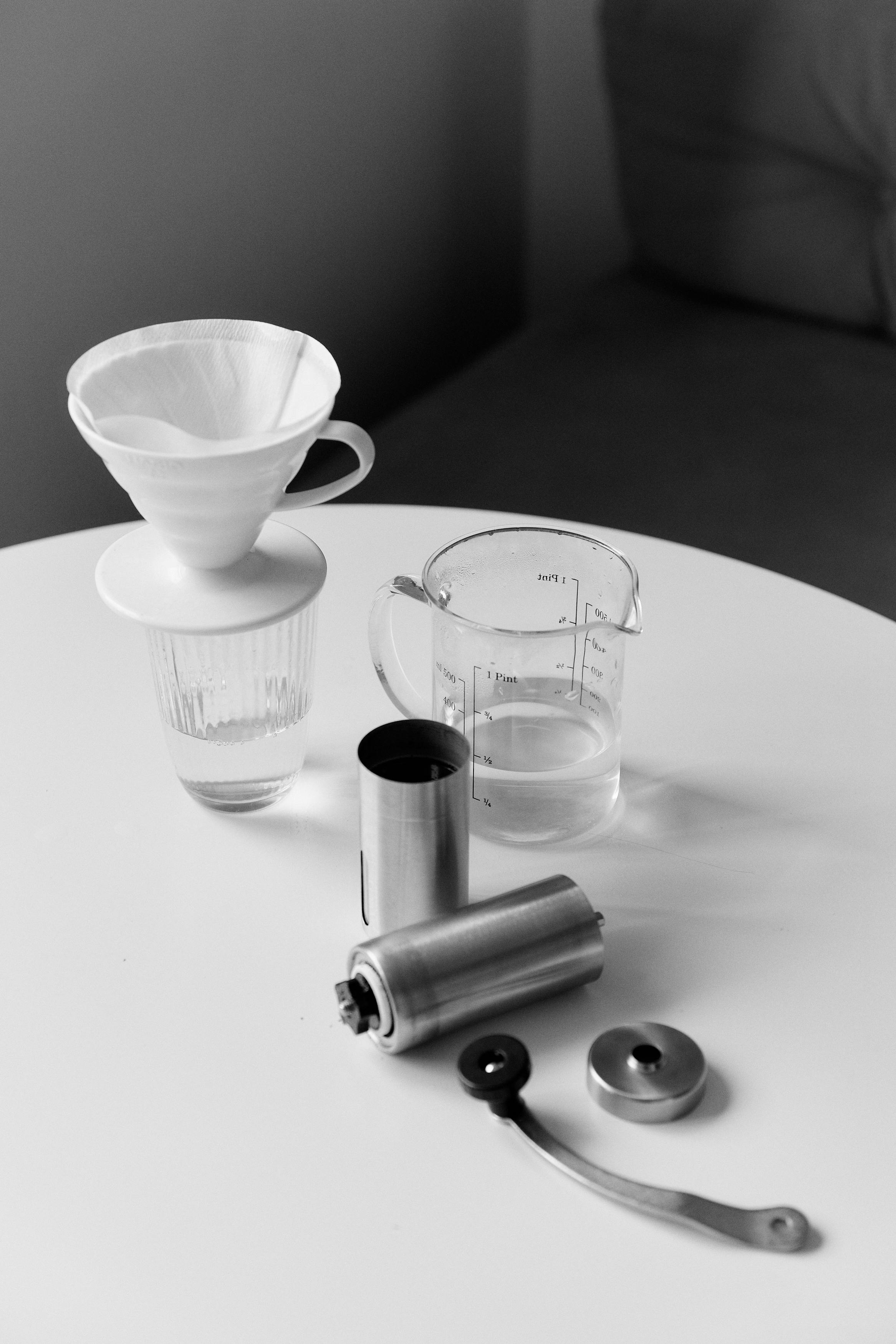 Grayscale image of coffee dripper, grinder, and pitcher on a white table.