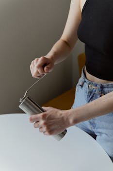 A woman in a black top and jeans hand-grinding coffee beans with a manual grinder indoors.