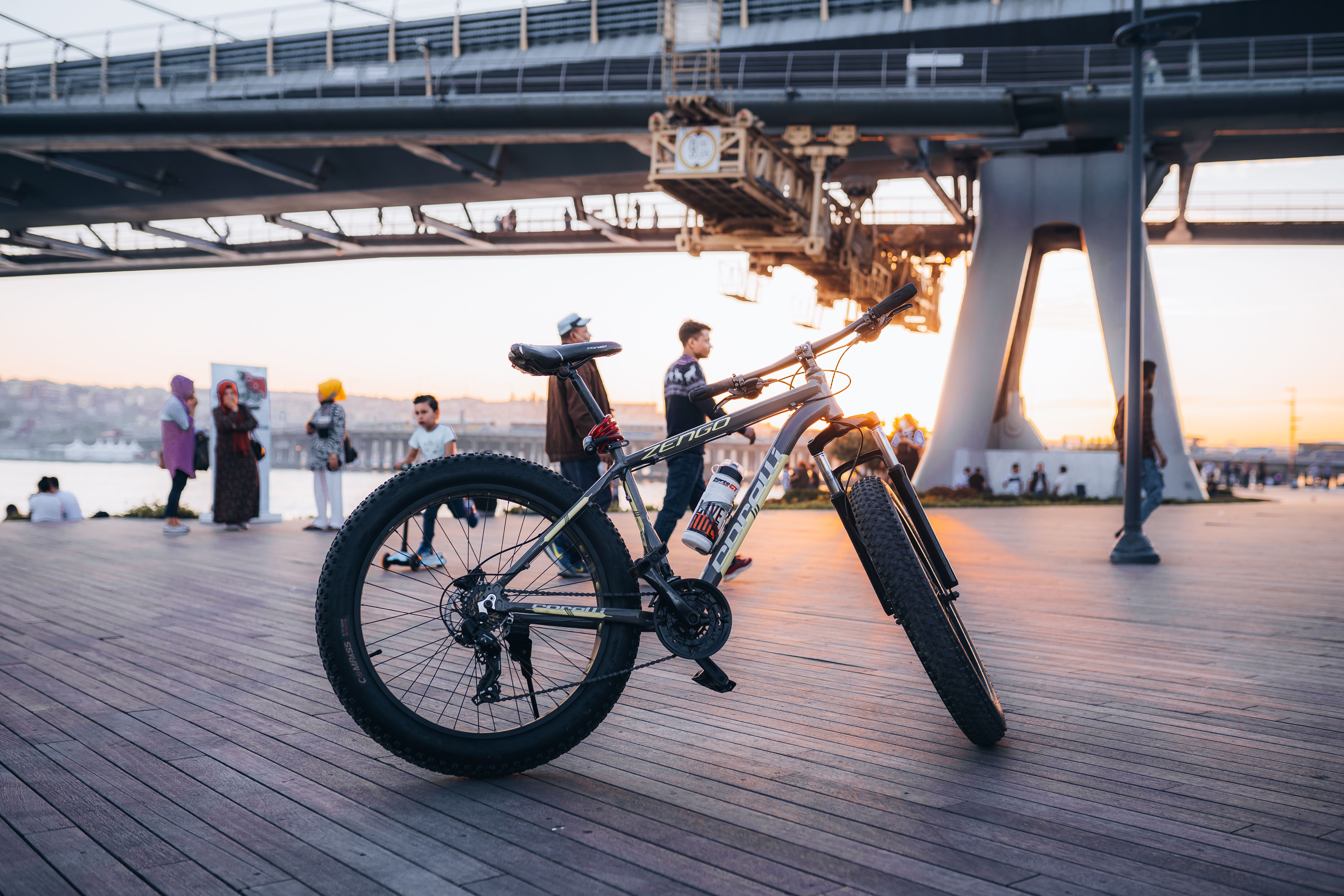 Bicycle parked on a wooden surface near a bridge during sunset with people in the background.