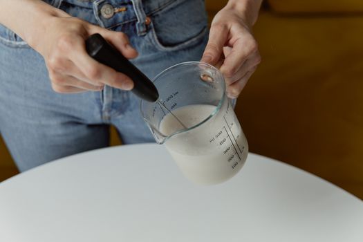 A person frothing milk with a handheld whisk in a transparent measuring cup, showcasing food preparation.