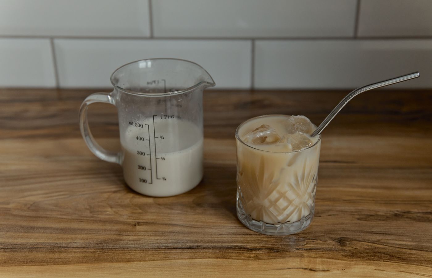 Iced coffee latte in crystal glass with metal straw next to measuring cup on wooden surface.