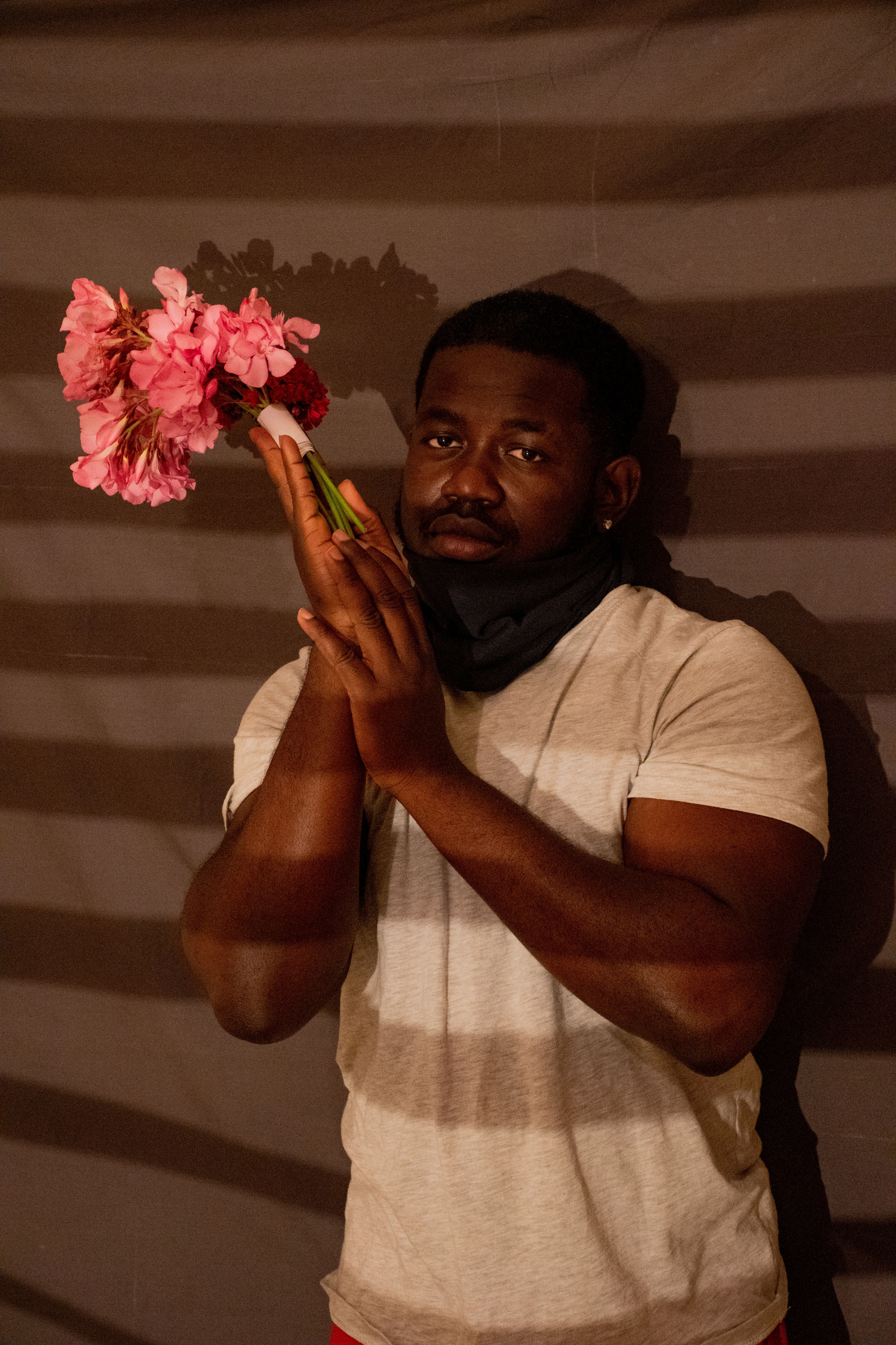 An adult man stands indoors holding vibrant pink flowers against a shadowed wall.