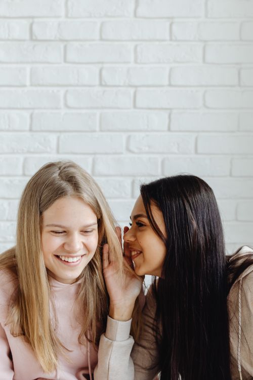 Free Two teenage girls whispering and smiling against a white brick wall, showcasing friendship and youthful joy. Stock Photo