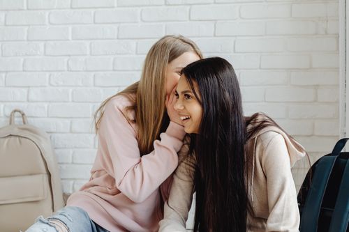 Free Two women in casual attire whisper and laugh against a white brick wall. Stock Photo