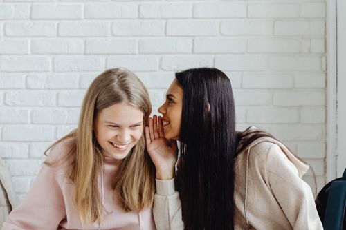 Free Two young women sharing a secret and laughing, dressed casually, indoors. Stock Photo