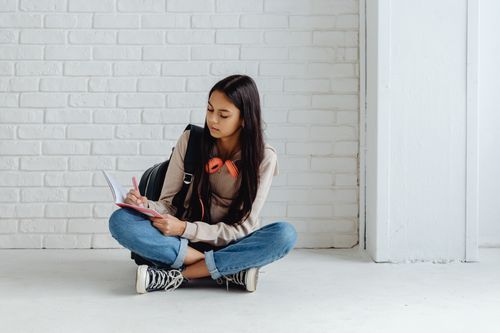 Free A young woman, wearing headphones and backpack, writes in a notebook while sitting on the floor in a bright, modern room. Stock Photo