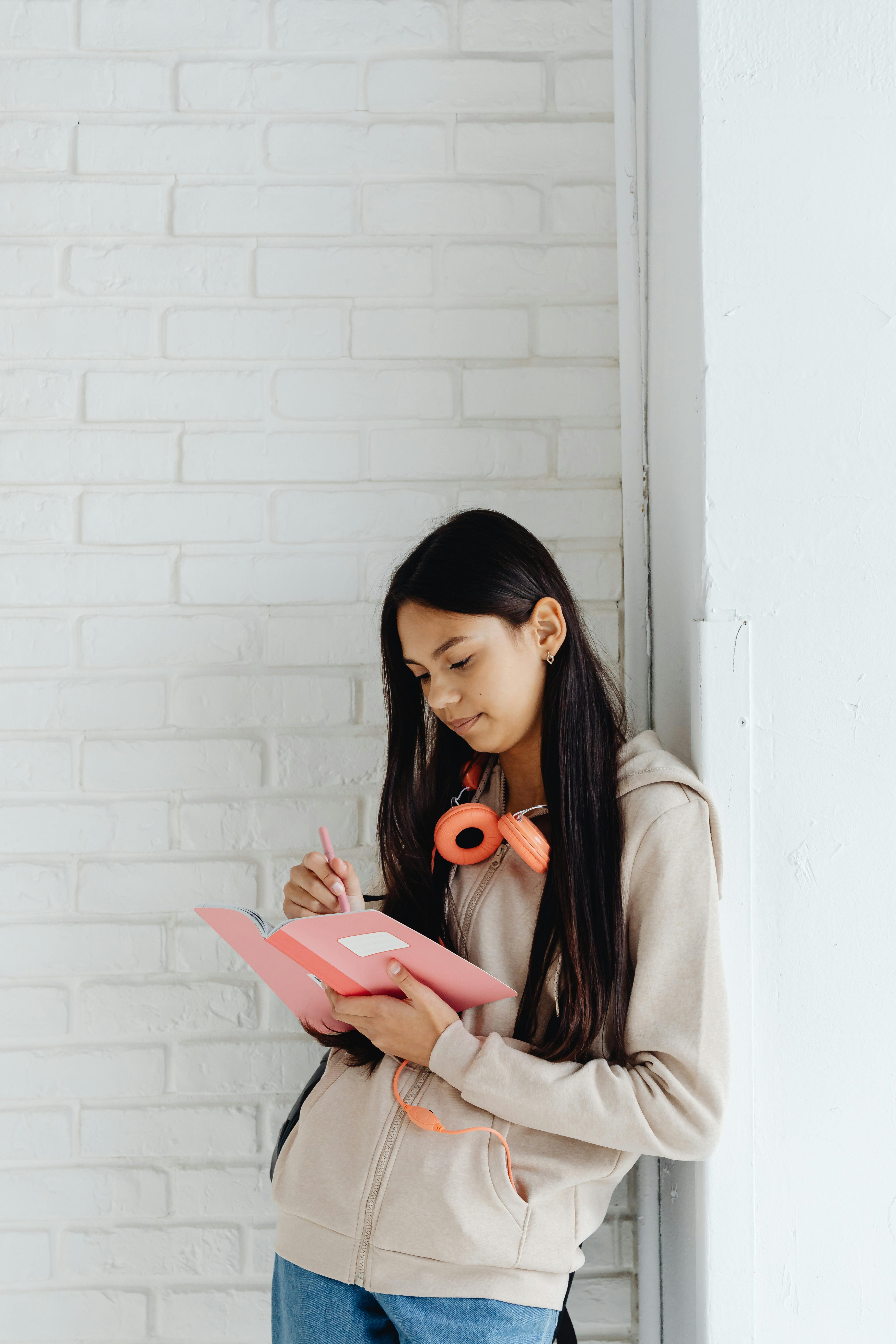 A girl with long, black hair writes in a pink journal. She has orange headphones around her neck, and is wearing a beige hoodie, jeans, and Converse. She is leaning against a white brick wall.