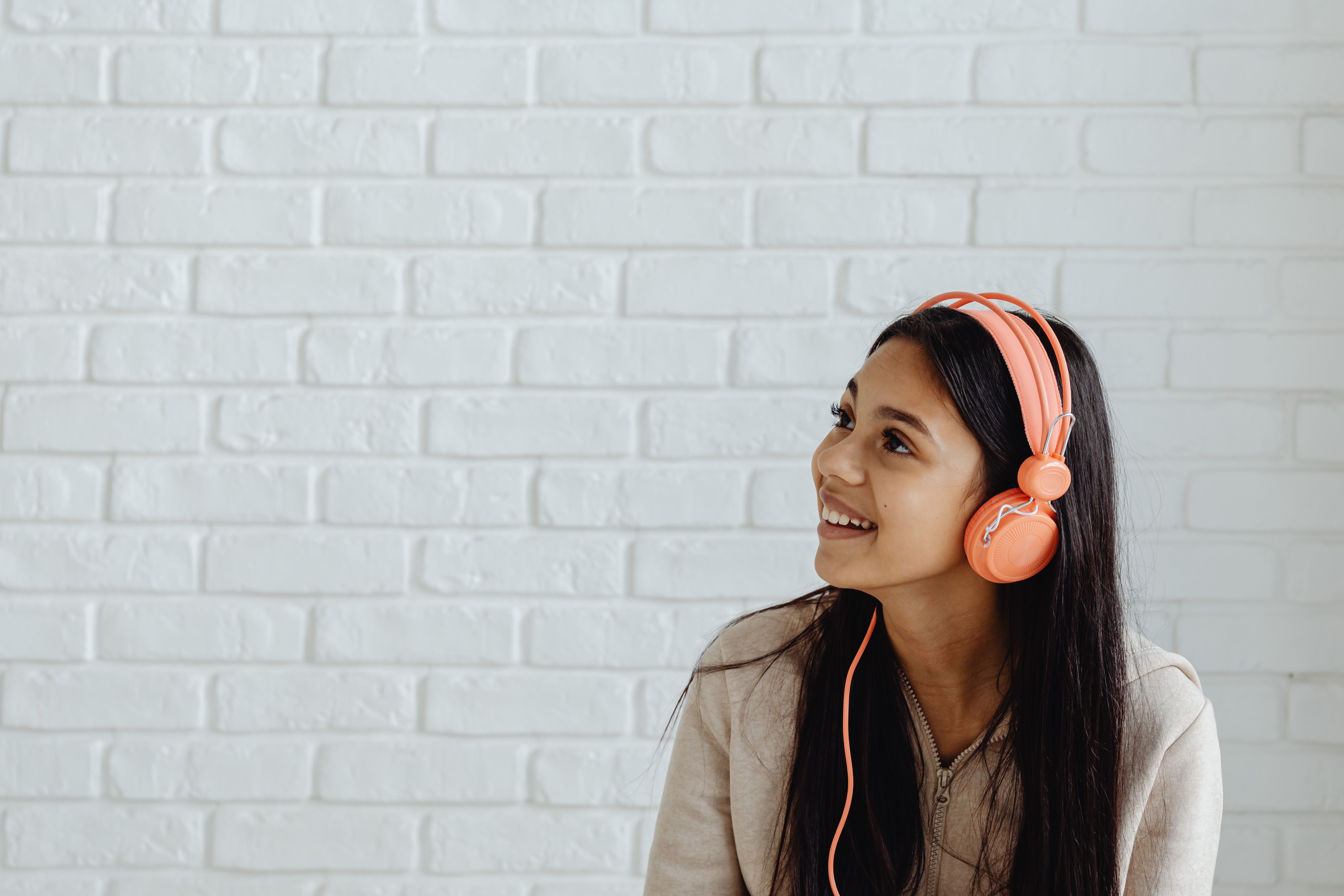 Smiling teenage girl wearing headphones against a white brick wall, enjoying music indoors.