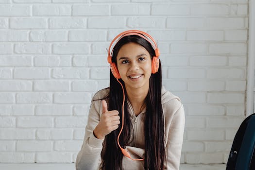 Smiling teenage girl sitting indoors, wearing headphones and giving thumbs up.