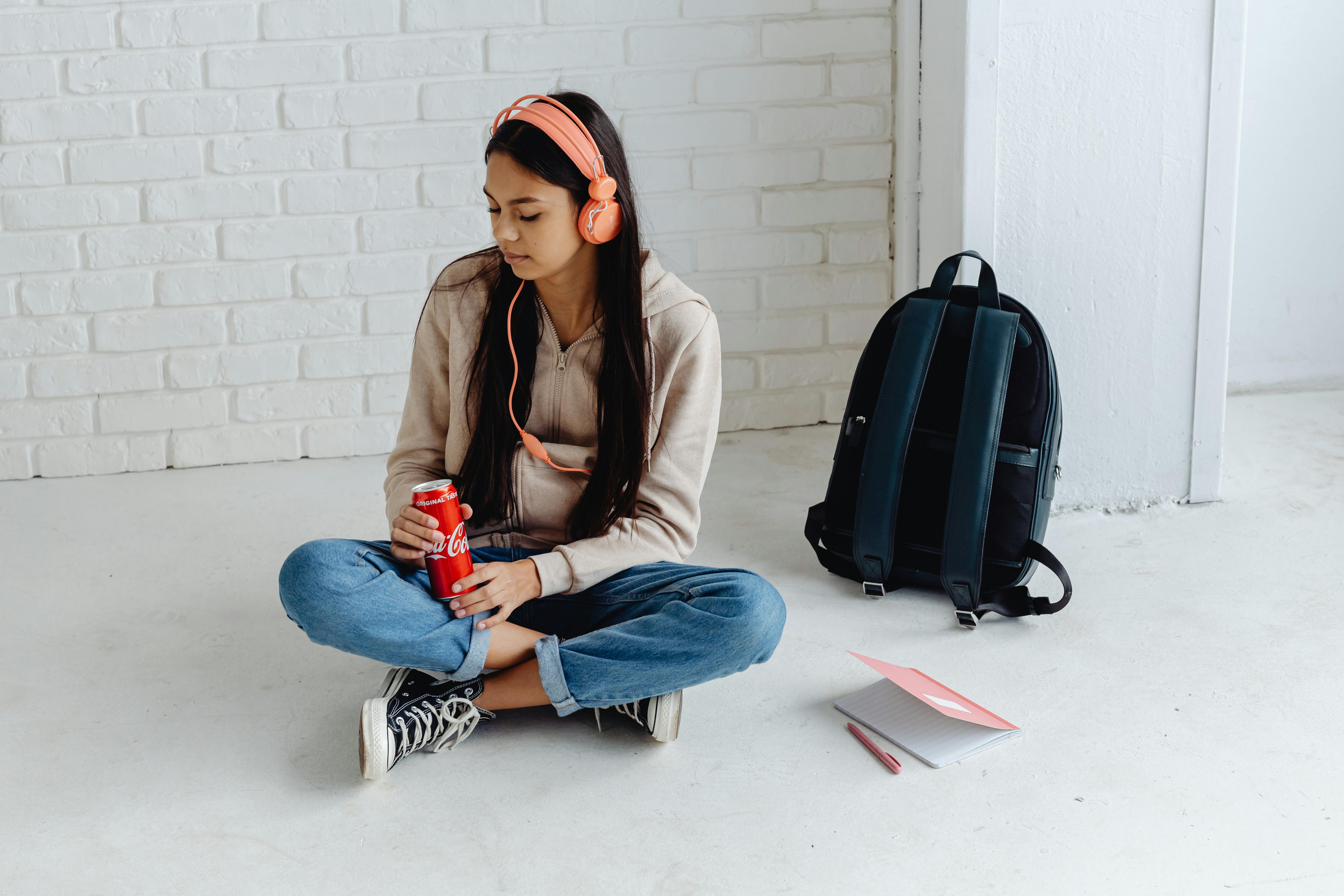 Teen girl with headphones, sitting in corridor, enjoying a drink in a relaxed atmosphere.