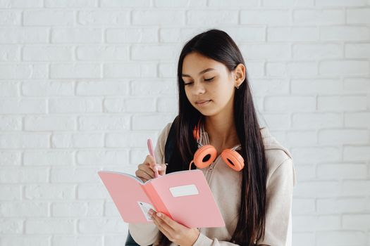 Teen girl writing notes in a pink notebook with headphones. Indoors setting.