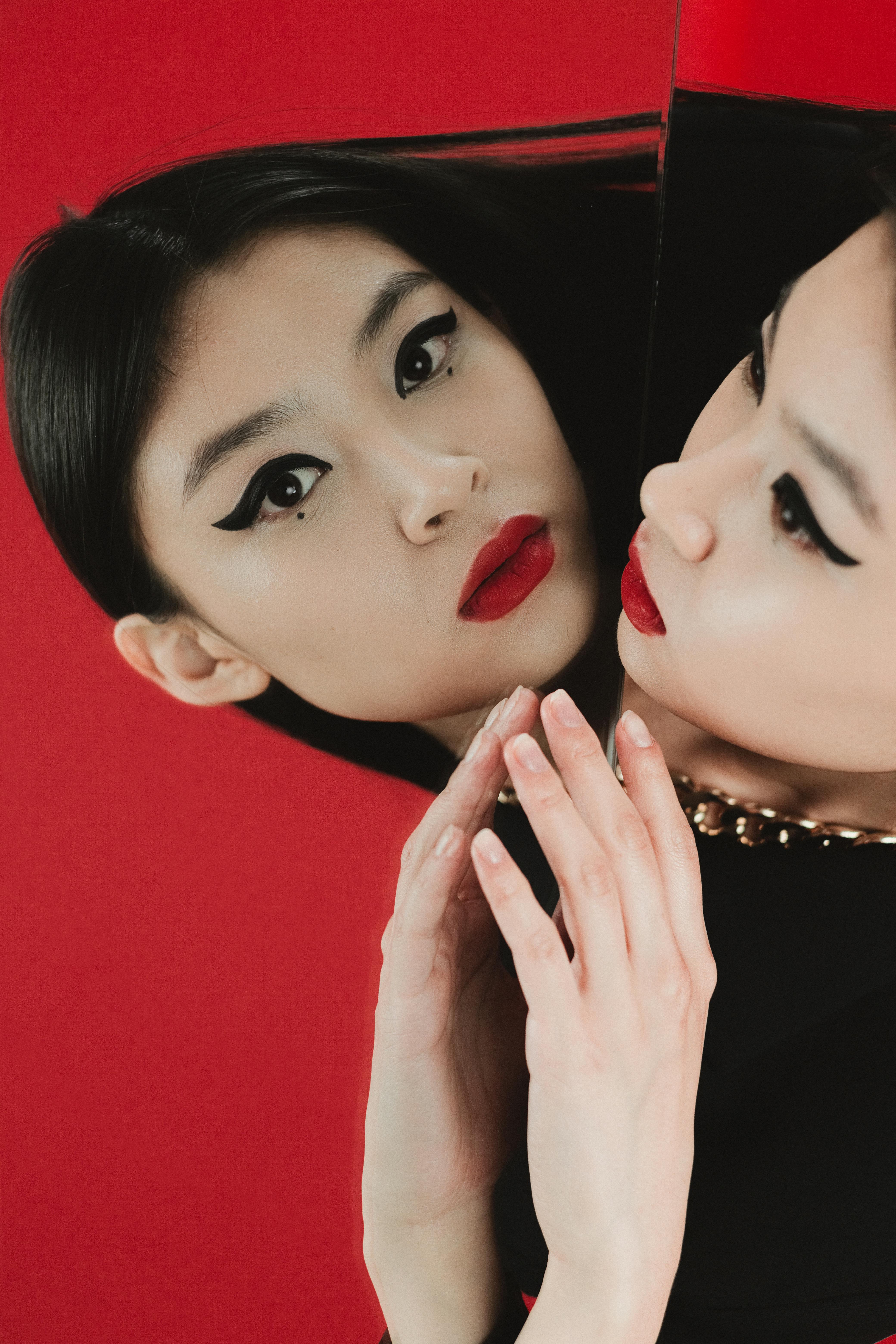 Stylish portrait of a woman with bold makeup and mirror reflection against a red background.