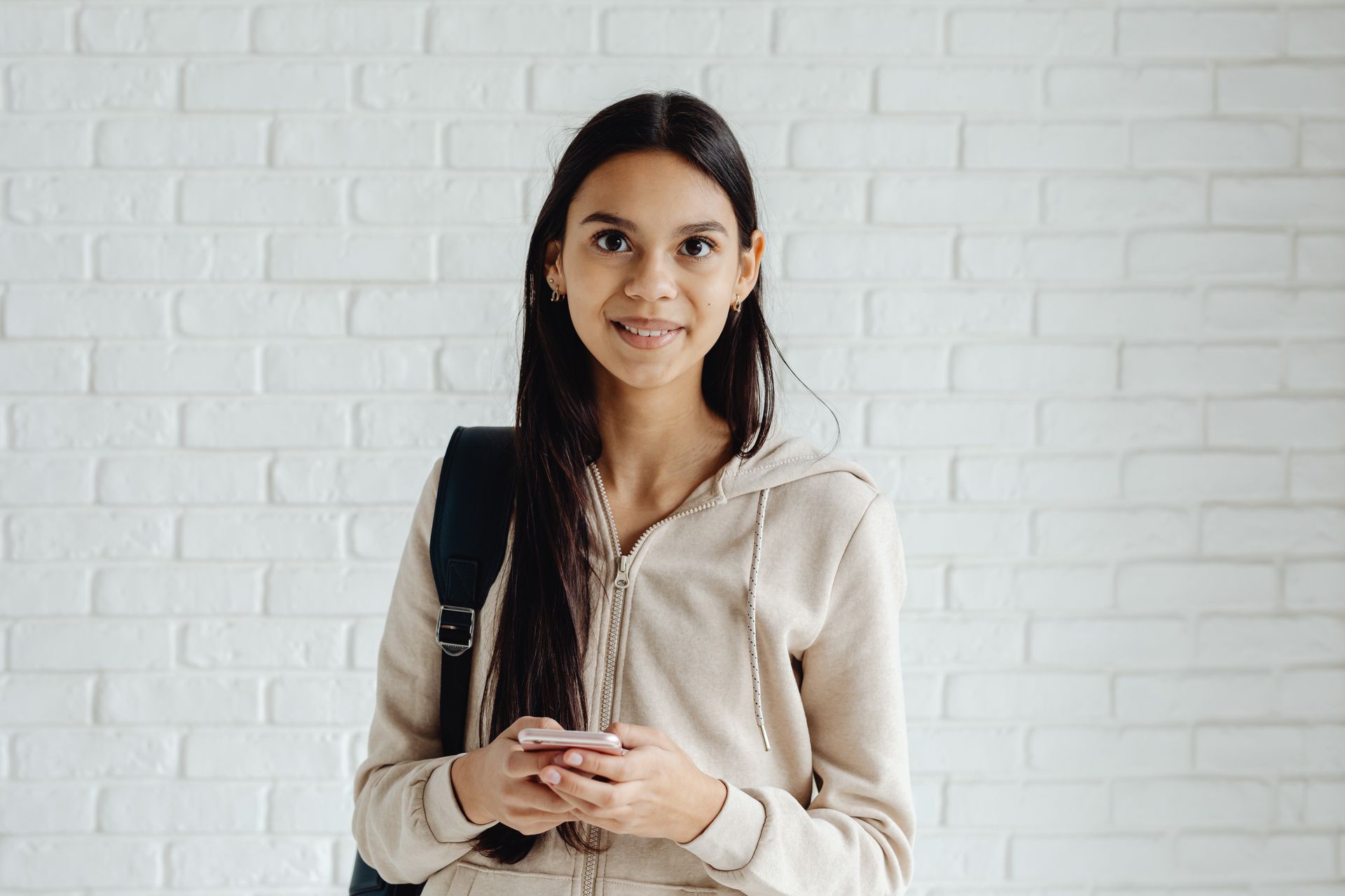Smiling young woman with long hair holding a phone in front of a white brick wall.