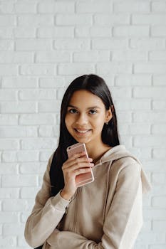 Young woman in beige hoodie smiling while holding smartphone in front of white brick wall.