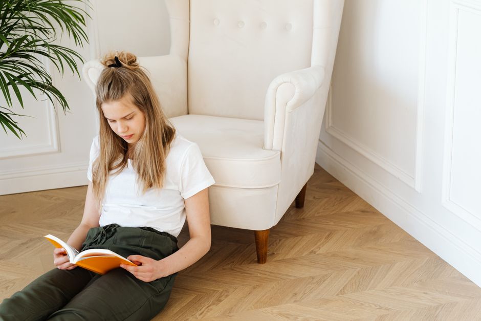 Teen girl reading a book beside a white armchair in a sunlit modern living room.