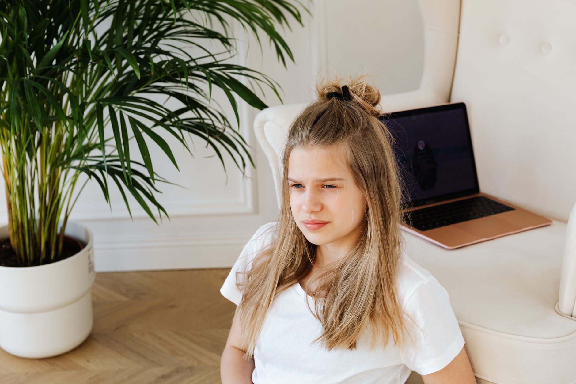 Junge blonde Frau im weißen T-Shirt sitzt neben einem Sofa mit aufgeklapptem Laptop