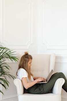 Teenage girl comfortably sitting in an armchair, focused on her laptop, studying indoors.