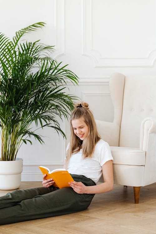 Free A Young Woman Sitting on the Wooden Floor while Reading Book Stock Photo