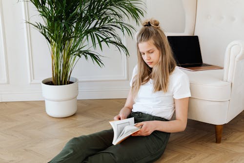 A Young Woman in White Shirt Sitting on the Floor while Busy Reading Book