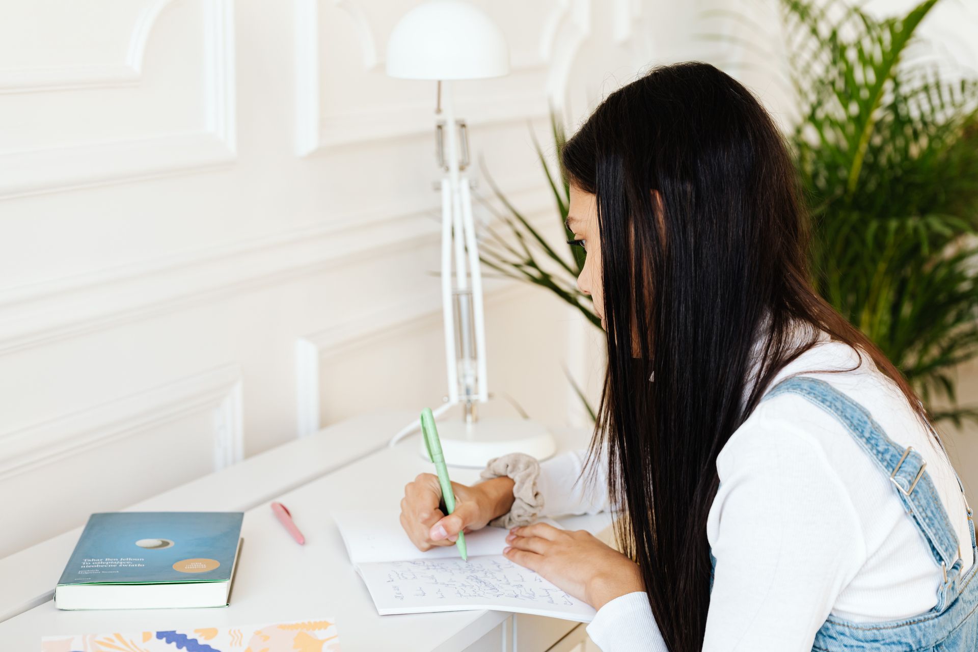 Teen girl writes in a notebook at a white desk, with books and plants nearby, under a lamp.
