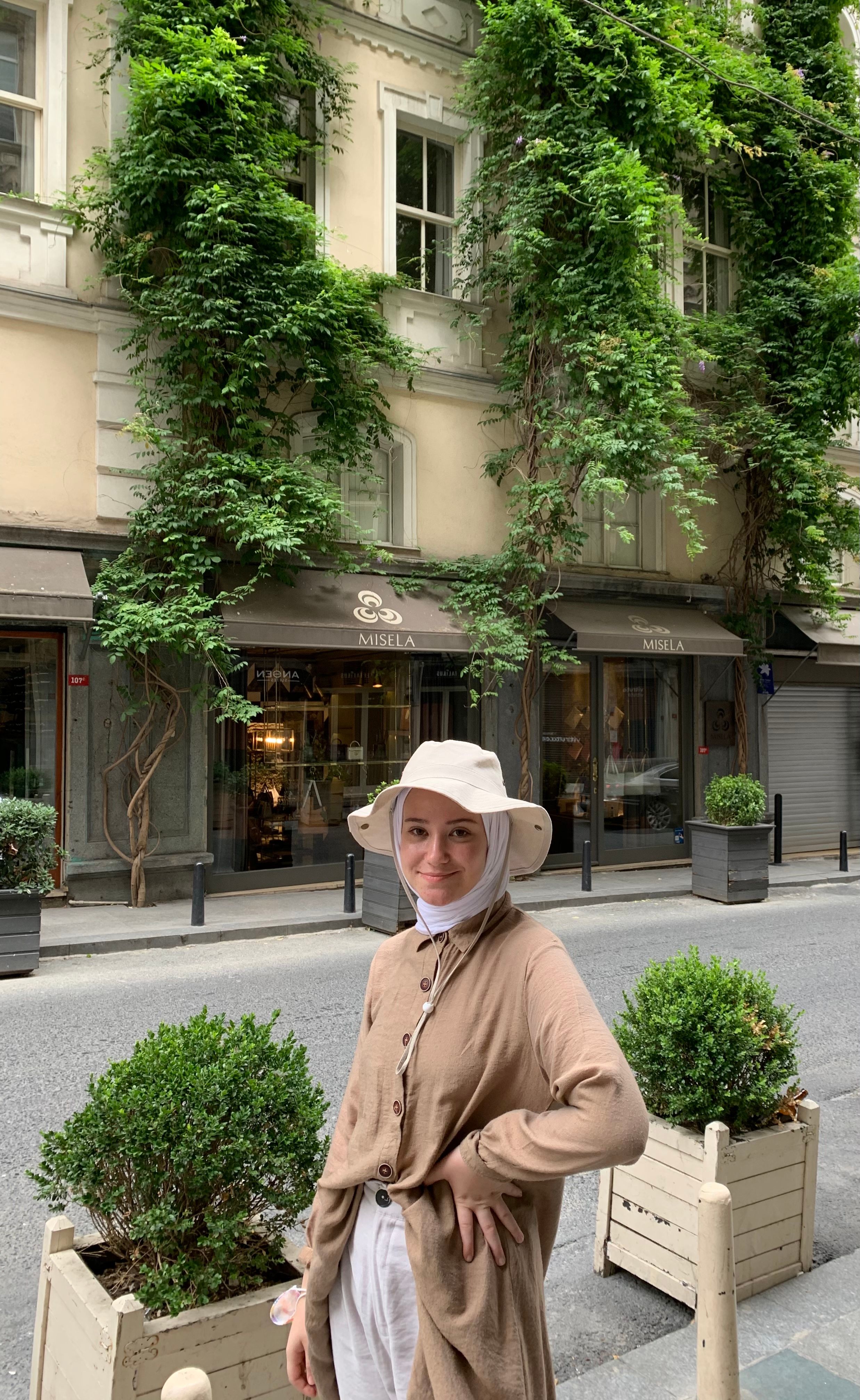 Woman in a hijab and hat smiling on an urban street in Istanbul, Türkiye, with greenery and buildings.